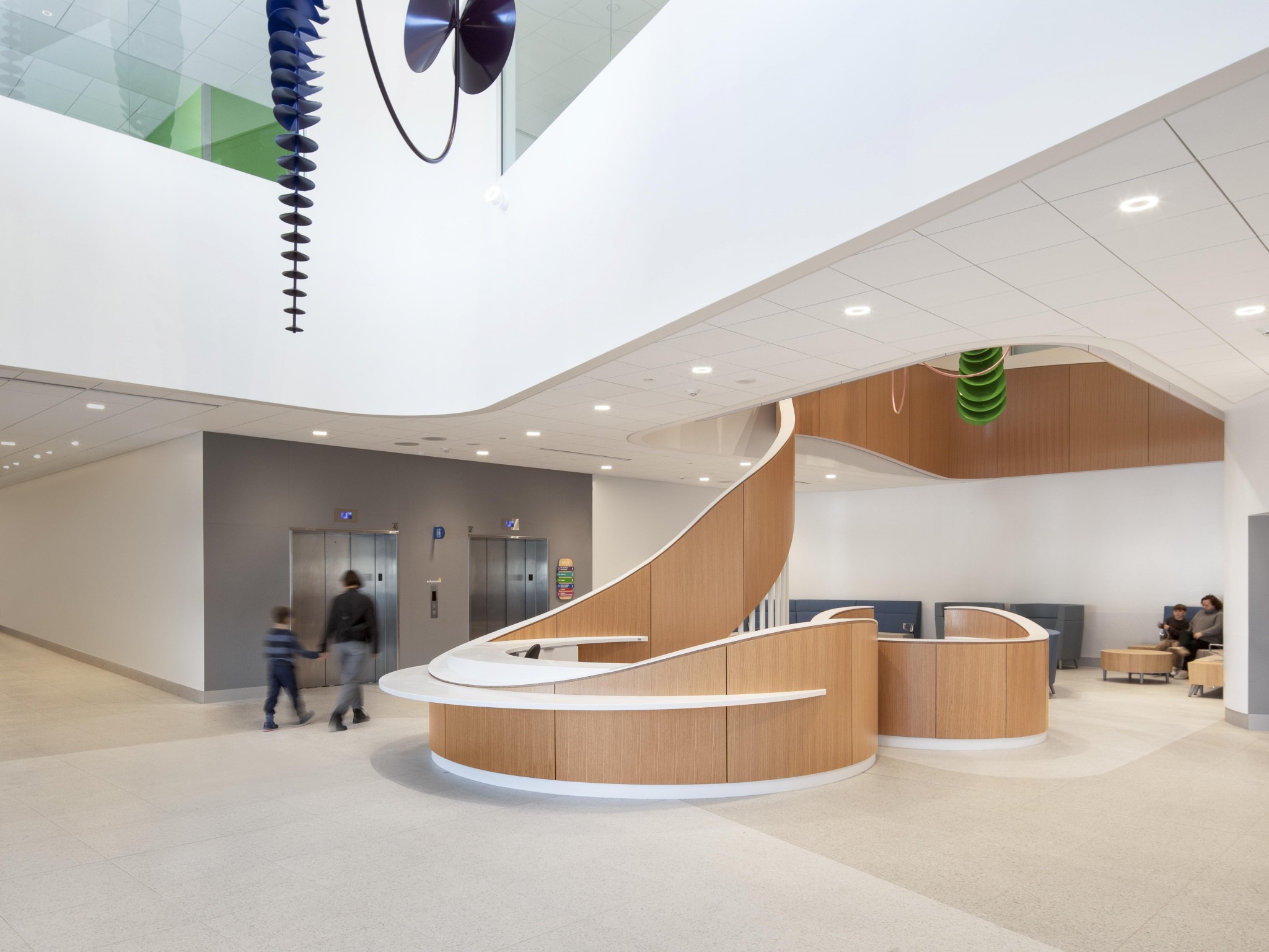 Modern, open lobby with wooden curved reception desk, spiral staircase, elevators, and people sitting and walking.