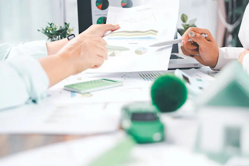 Two people reviewing financial charts and graphs at a desk with a calculator, laptop, and small model house in the foreground.