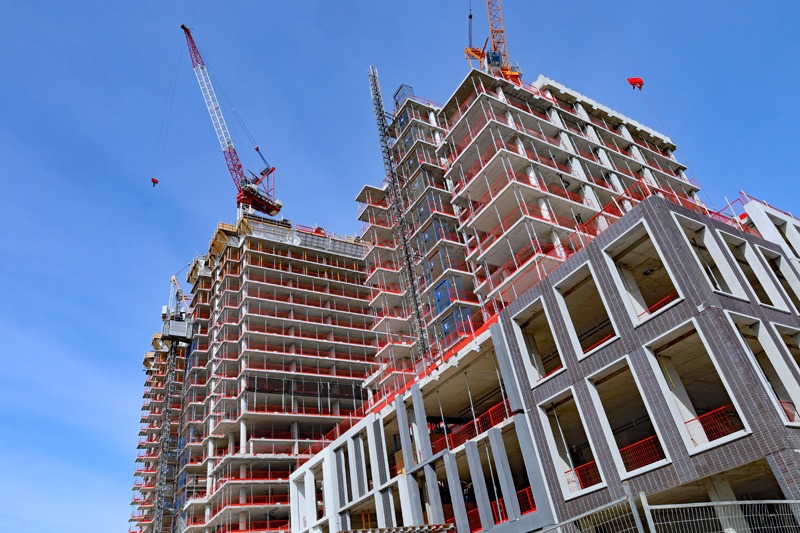 Two high-rise buildings under construction with cranes set against a clear blue sky. Scaffolding and red safety netting are visible.