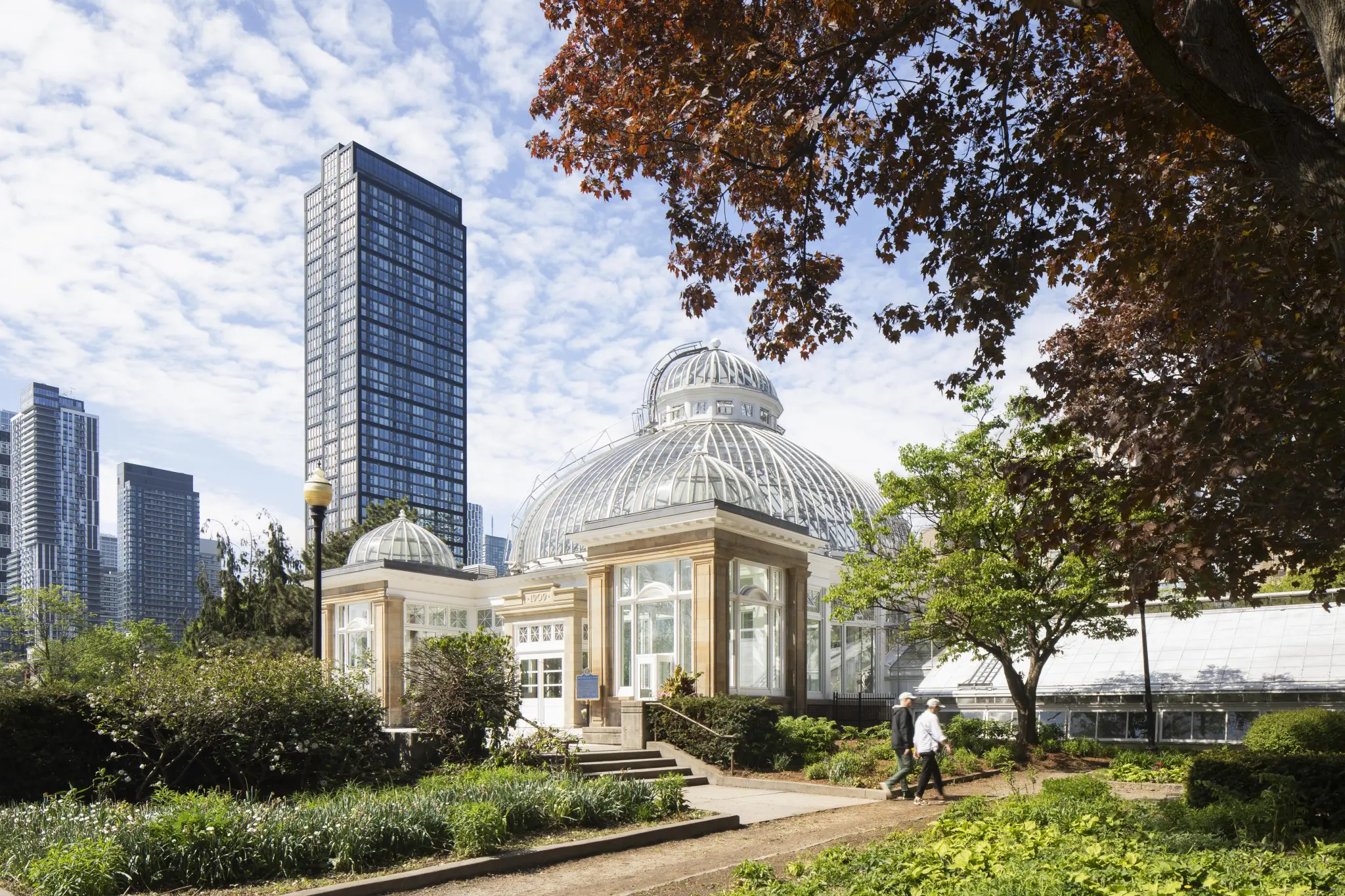 A charming conservatory with a domed glass roof is surrounded by lush gardens and tall city skyscrapers. Two people walk along a path under a sky dappled with clouds.
