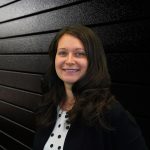 Smiling woman with long dark hair wearing a polka dot shirt and black blazer, standing against a textured black wall. The tone is professional and approachable.