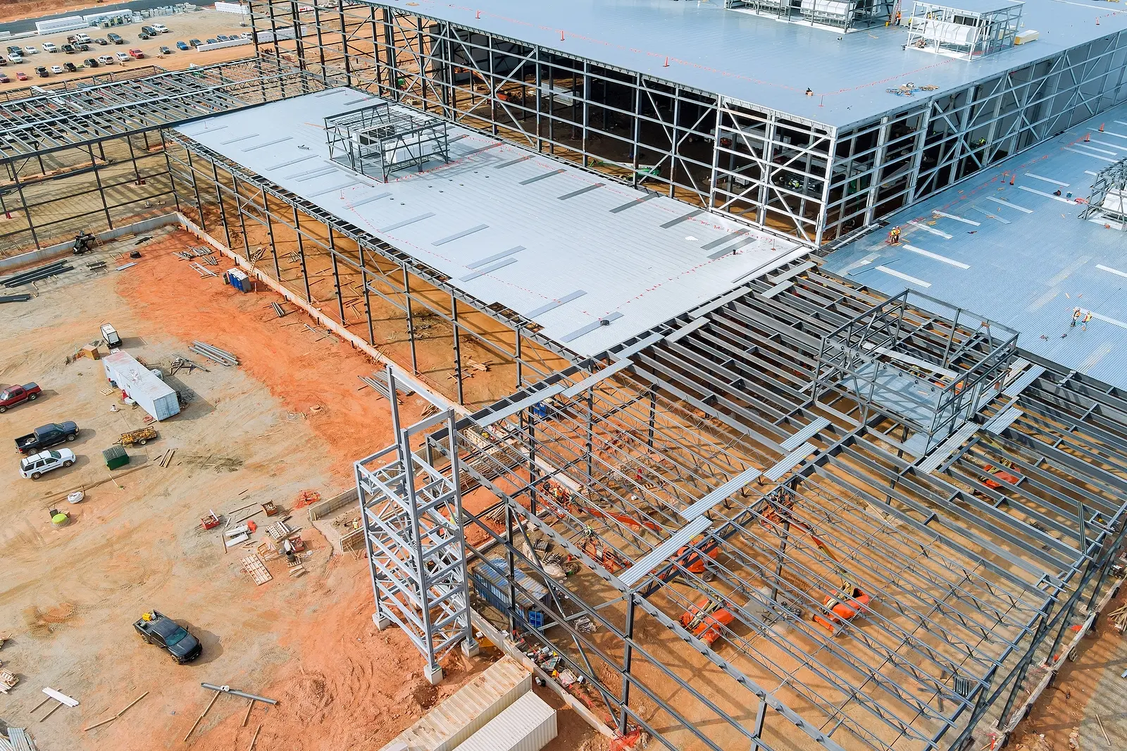 Aerial view of a large industrial building under construction. Steel framework and metal roofing dominate the site, with scattered construction vehicles and earthmoving equipment on red soil.