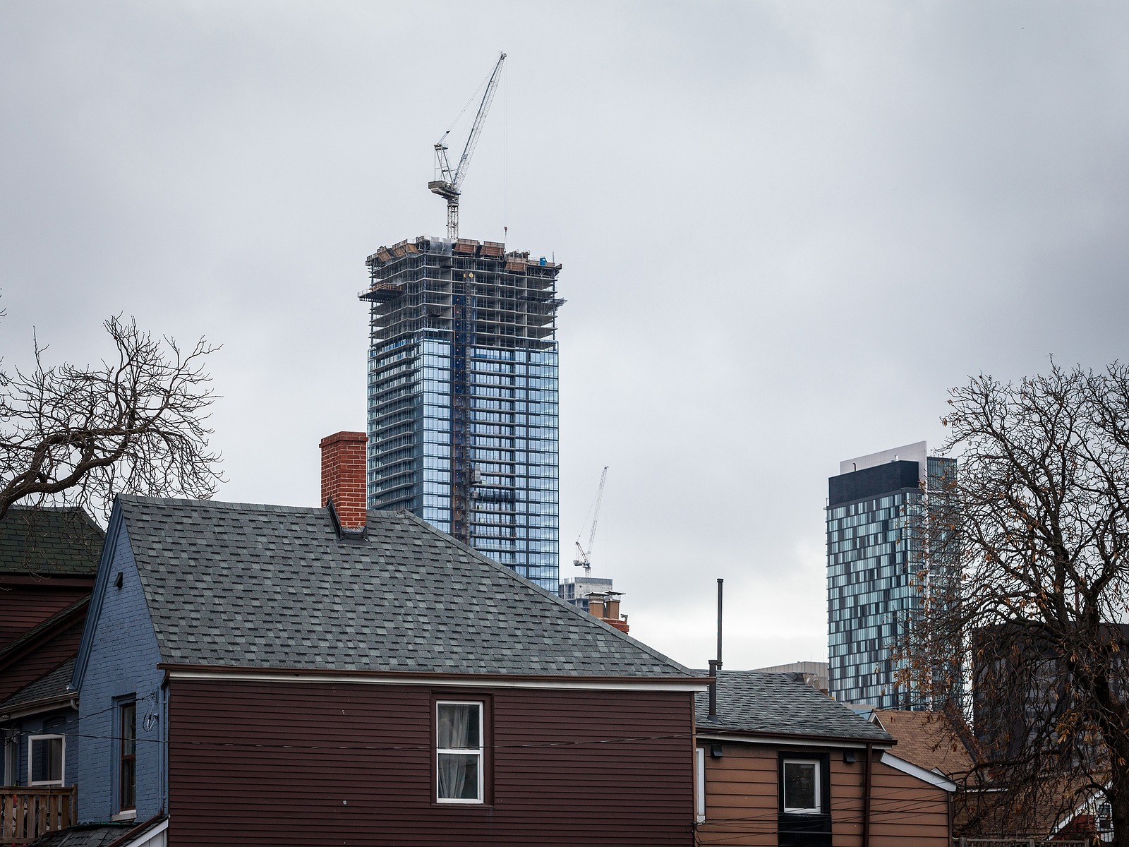 A towering modern skyscraper under construction, topped with a crane, contrasts against overcast skies and low-rise residential homes in the foreground.