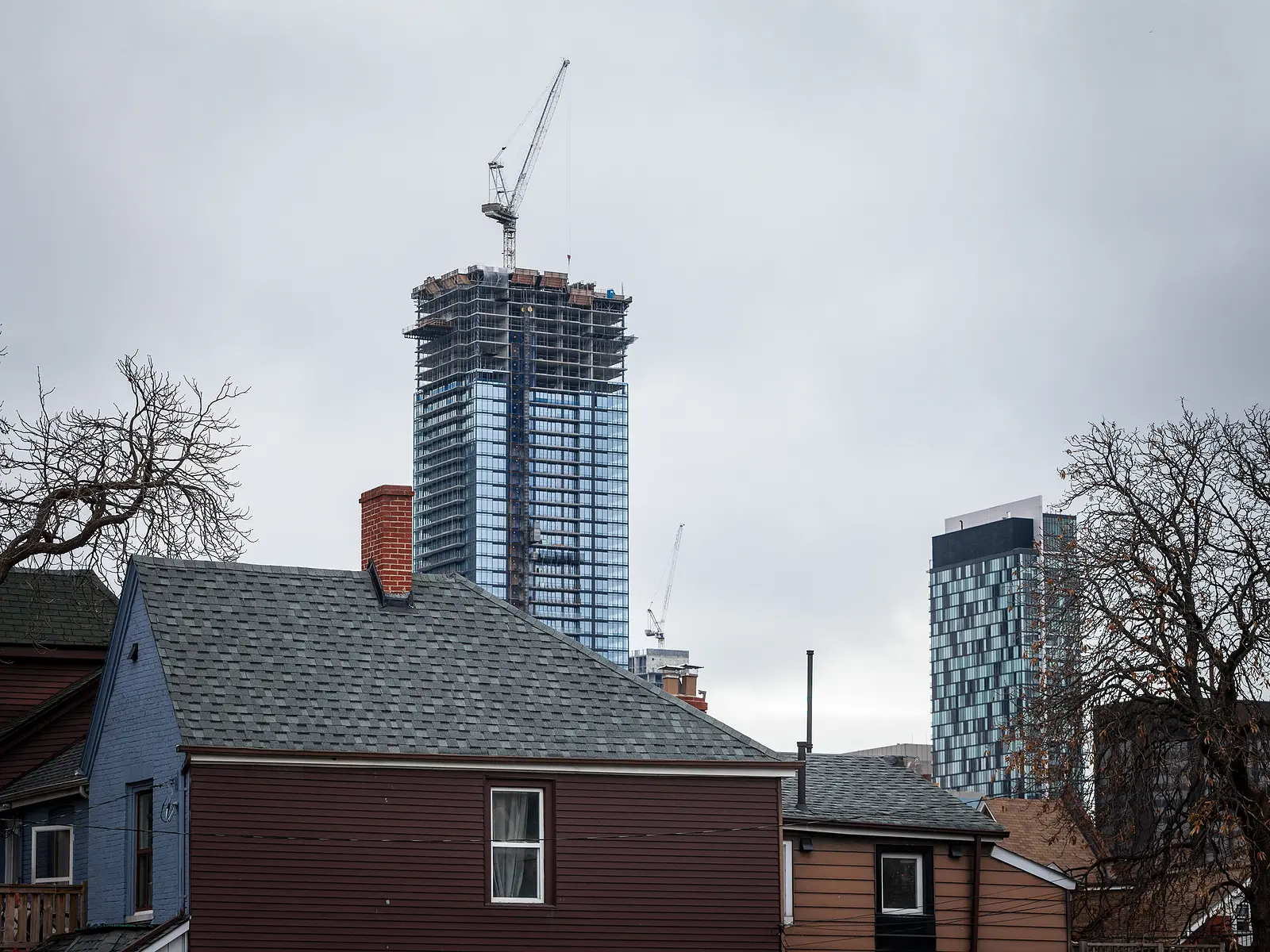A towering modern skyscraper under construction, topped with a crane, contrasts against overcast skies and low-rise residential homes in the foreground.