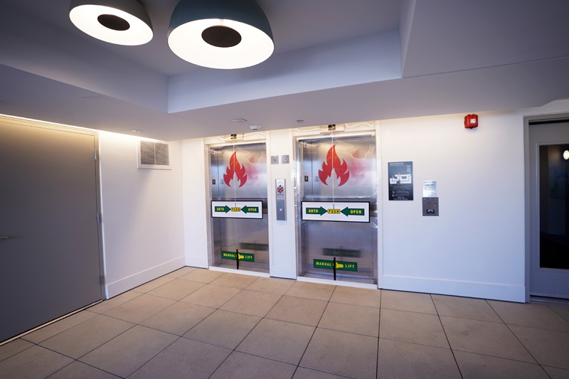 Modern building lobby with two stainless steel fire exit doors featuring red flame symbols. Bright overhead lighting, white walls, and tiled floor.