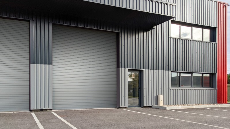 Modern industrial warehouse exterior with gray metal siding and large roller door. Adjacent are windows and a red accent stripe. The scene feels clean and functional.