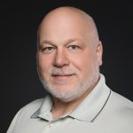 Bald man with a beard, wearing a light gray polo shirt featuring a logo. He smiles warmly against a dark background. Professional and approachable tone.