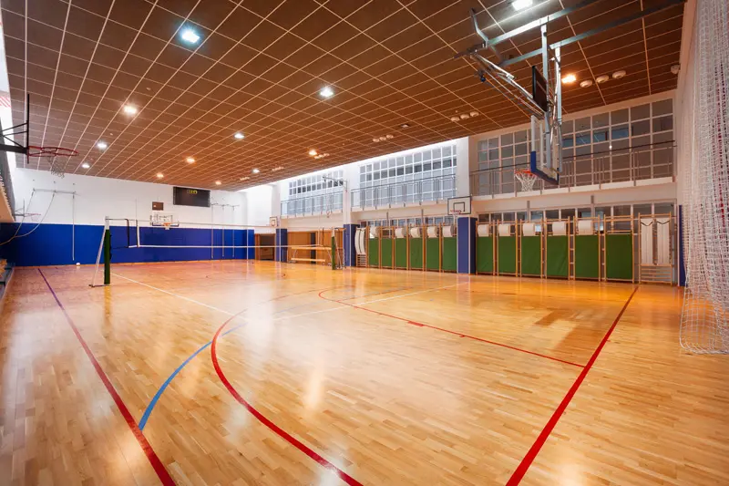 Spacious indoor gymnasium with polished wooden floor, basketball hoops, and volleyball nets. Bright lighting and clean, organized setup convey readiness.