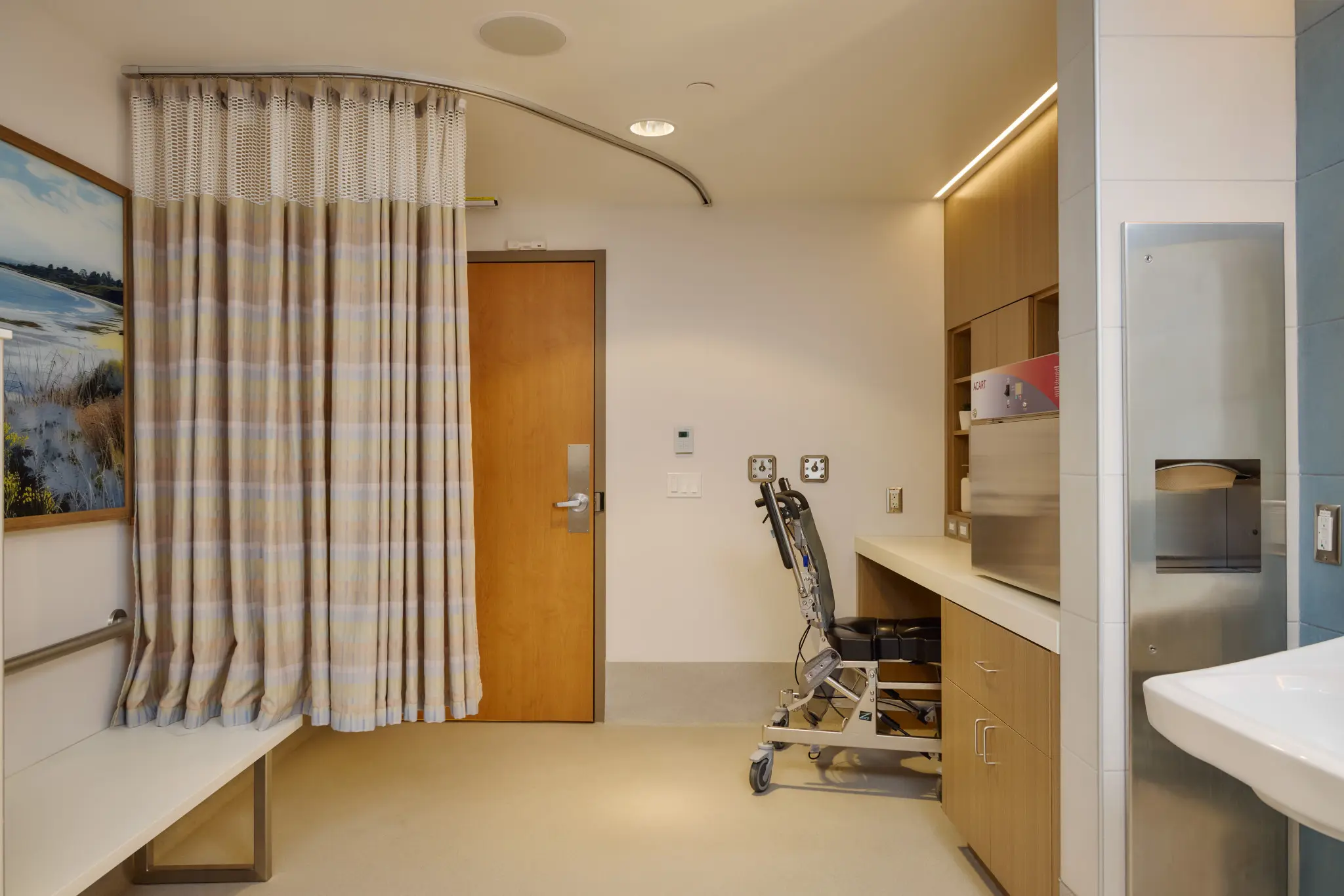 Hospital room with a neutral tone, featuring a striped privacy curtain, wooden cabinets, a medical chair, and a calm landscape painting.