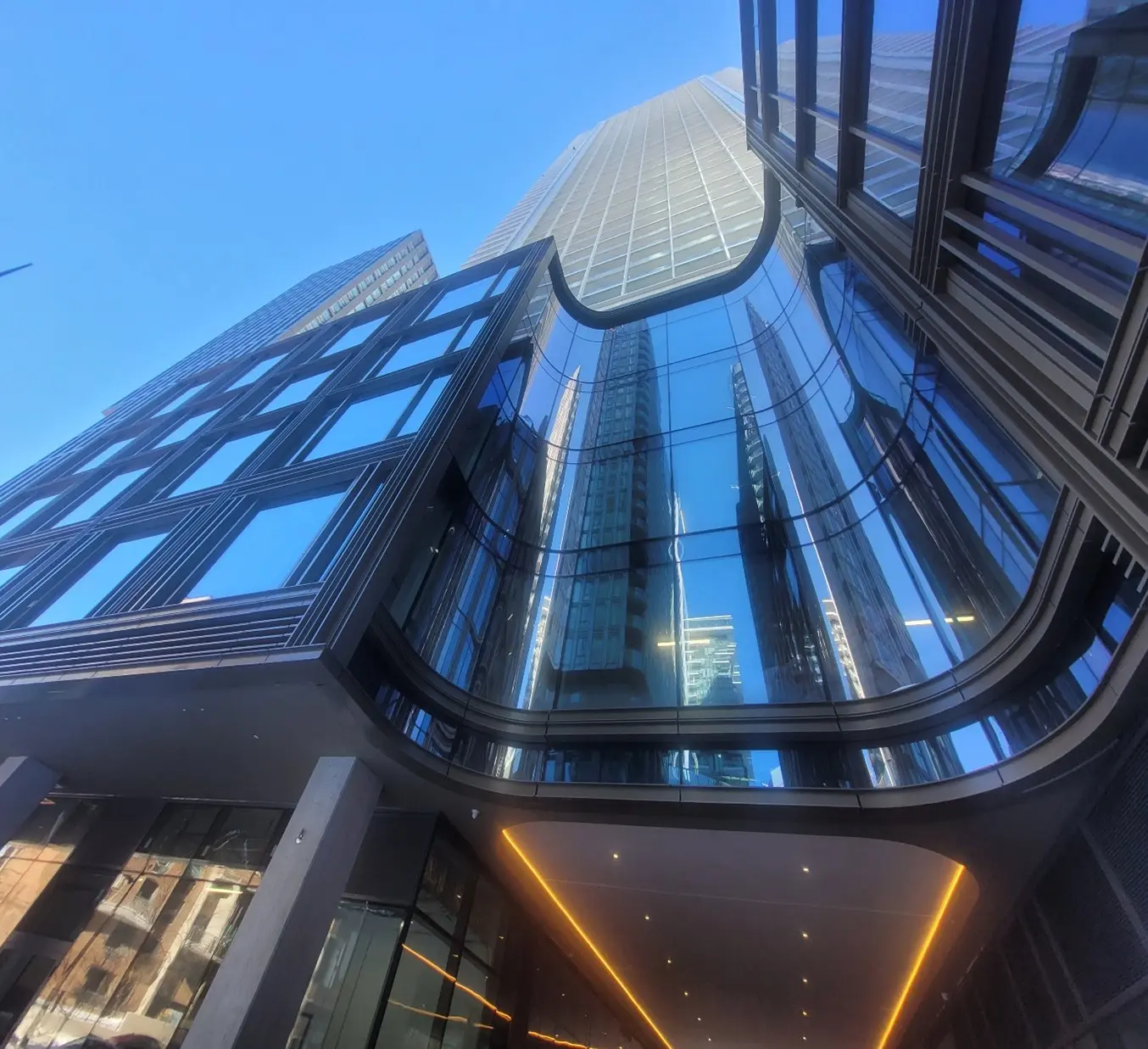 Upward view of a modern skyscraper with reflective glass facade, capturing the blue sky and other buildings. Sleek, futuristic design exudes elegance.