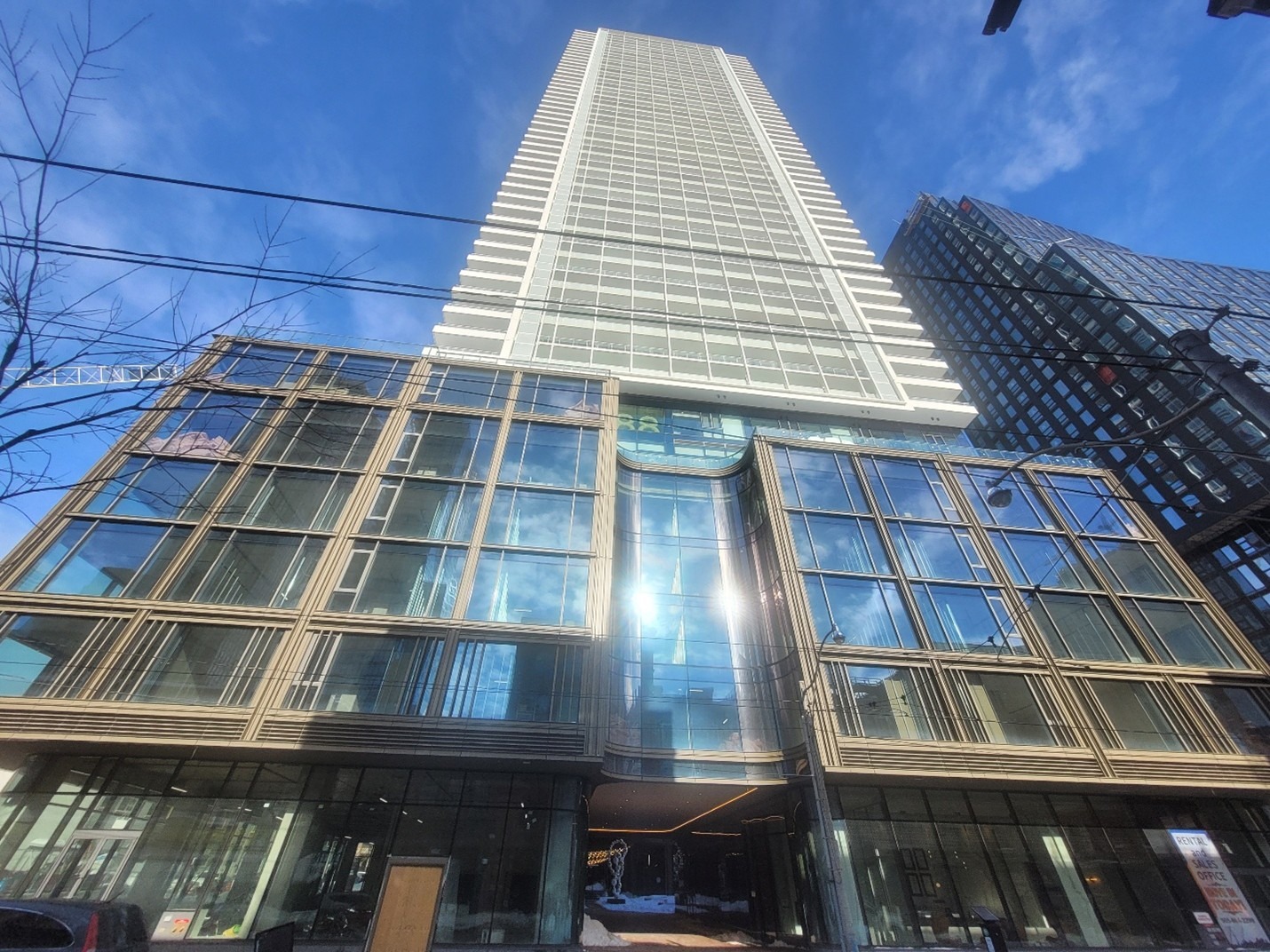 Tall, modern skyscraper under a blue sky, with reflective glass facade and grid-like design. Nearby buildings and bare tree branches surround it. Urban, imposing feel.