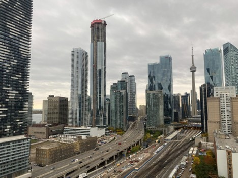 A cloudy skyline view of Toronto featuring modern skyscrapers, the CN Tower, and a network of rail tracks and highways in the foreground.