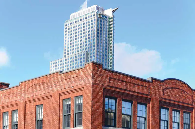 Red brick building with large windows in the foreground, contrasted by a modern, glass-covered skyscraper in the background against a clear sky.