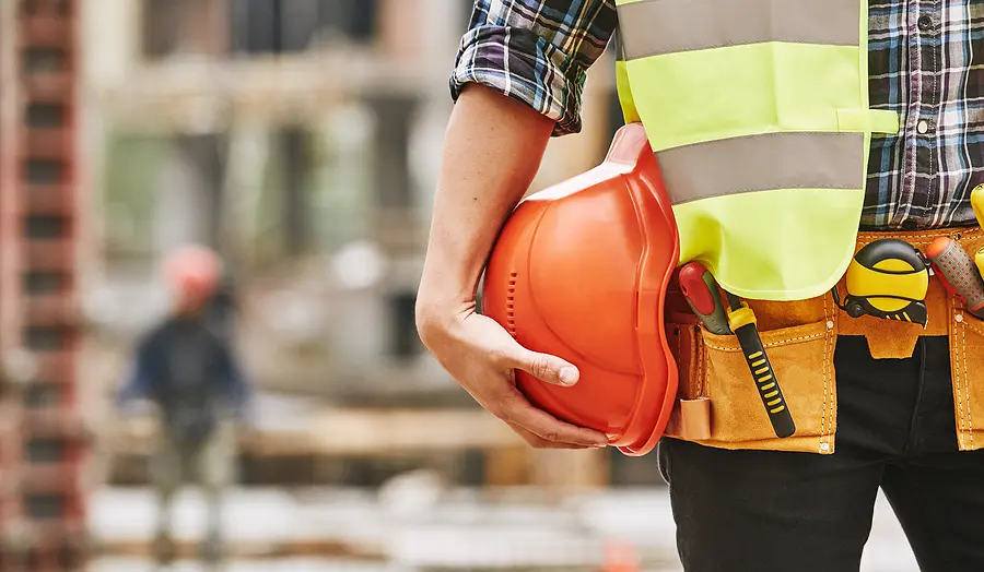 Cropped photo of male professional builder in working uniform with construction tools holding a safety red helmet while standing outdoor of construction site.