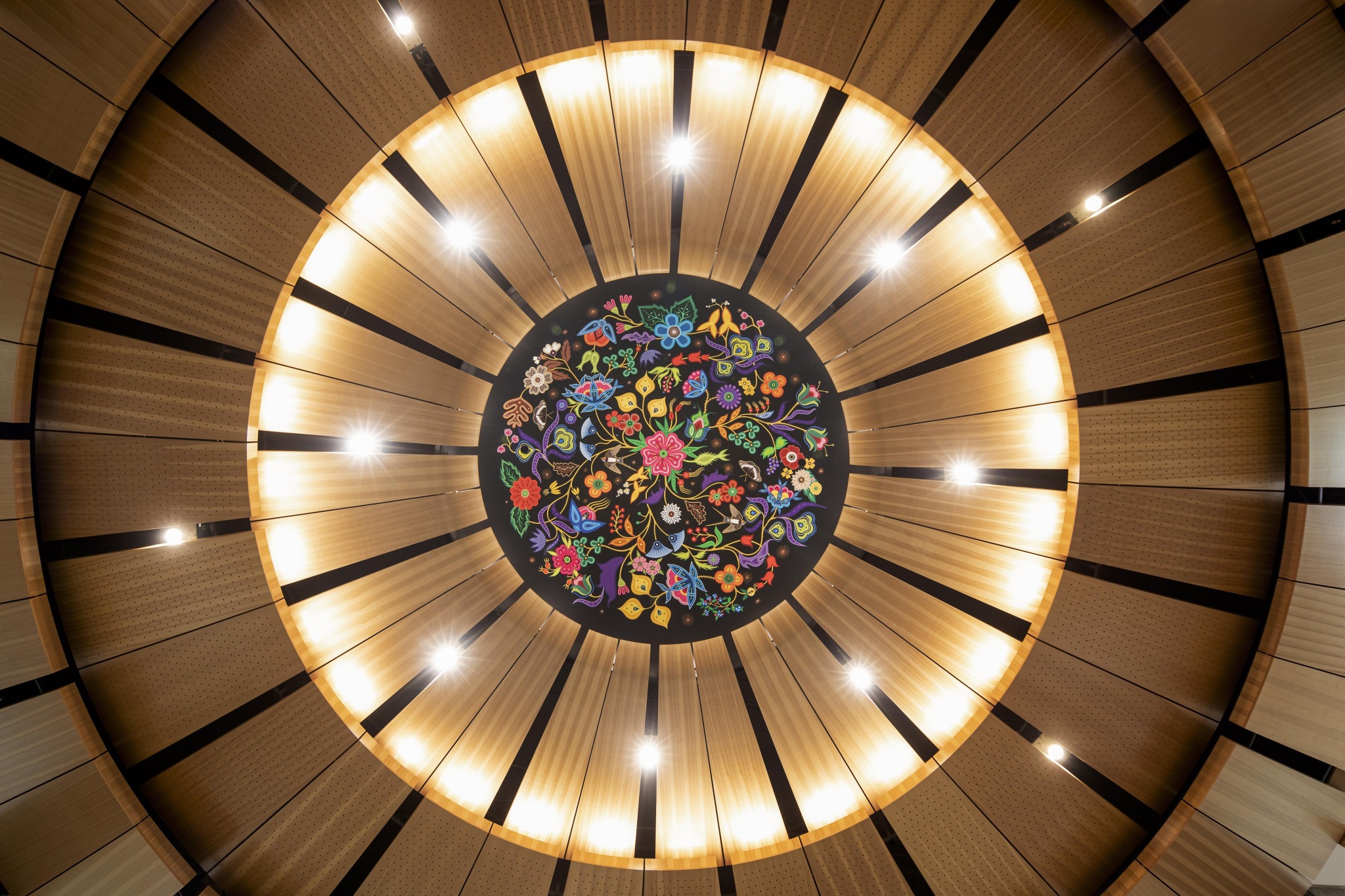 Circular wood ceiling with a central colorful floral pattern, illuminated by evenly spaced lights. The design conveys elegance and artistic creativity.
