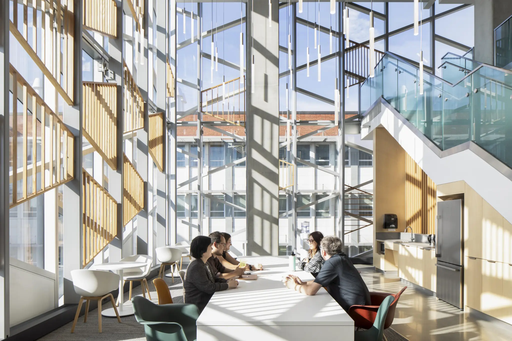 Spacious modern interior with high ceilings, large windows, and hanging pendant lights. Two people sit at a central table. Bright, airy atmosphere.