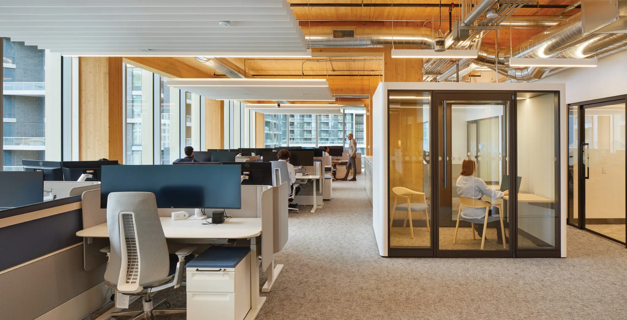 Modern office with open workstations and glass-walled meeting booths. Natural light, wooden accents, and carpeted floors create a calm, professional atmosphere.