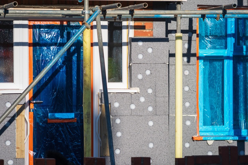Scaffolding surrounds a house facade with blue film-covered door and windows. The wall is gray with white dots, indicating ongoing construction work.