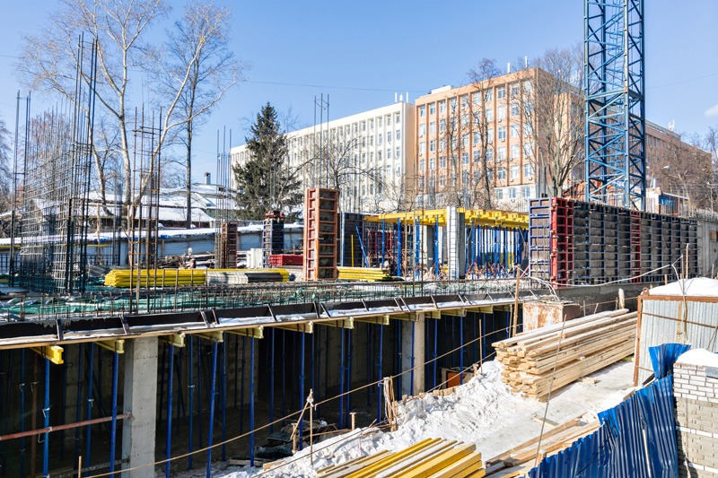 Construction site with steel frameworks and stacks of wooden beams under a clear blue sky. A crane and buildings can be seen in the background.