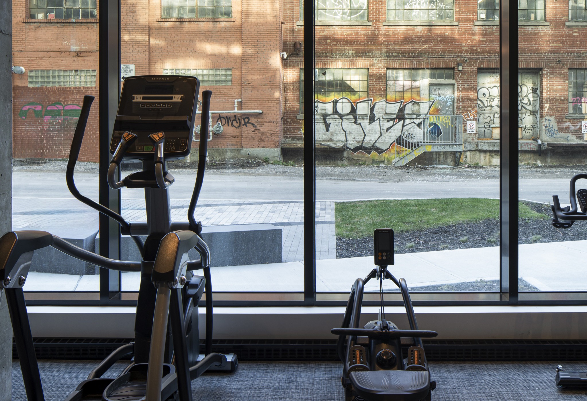 Gym interior with exercise equipment in the foreground, facing large windows. Outside, a brick wall with colorful graffiti is visible, creating an urban vibe.