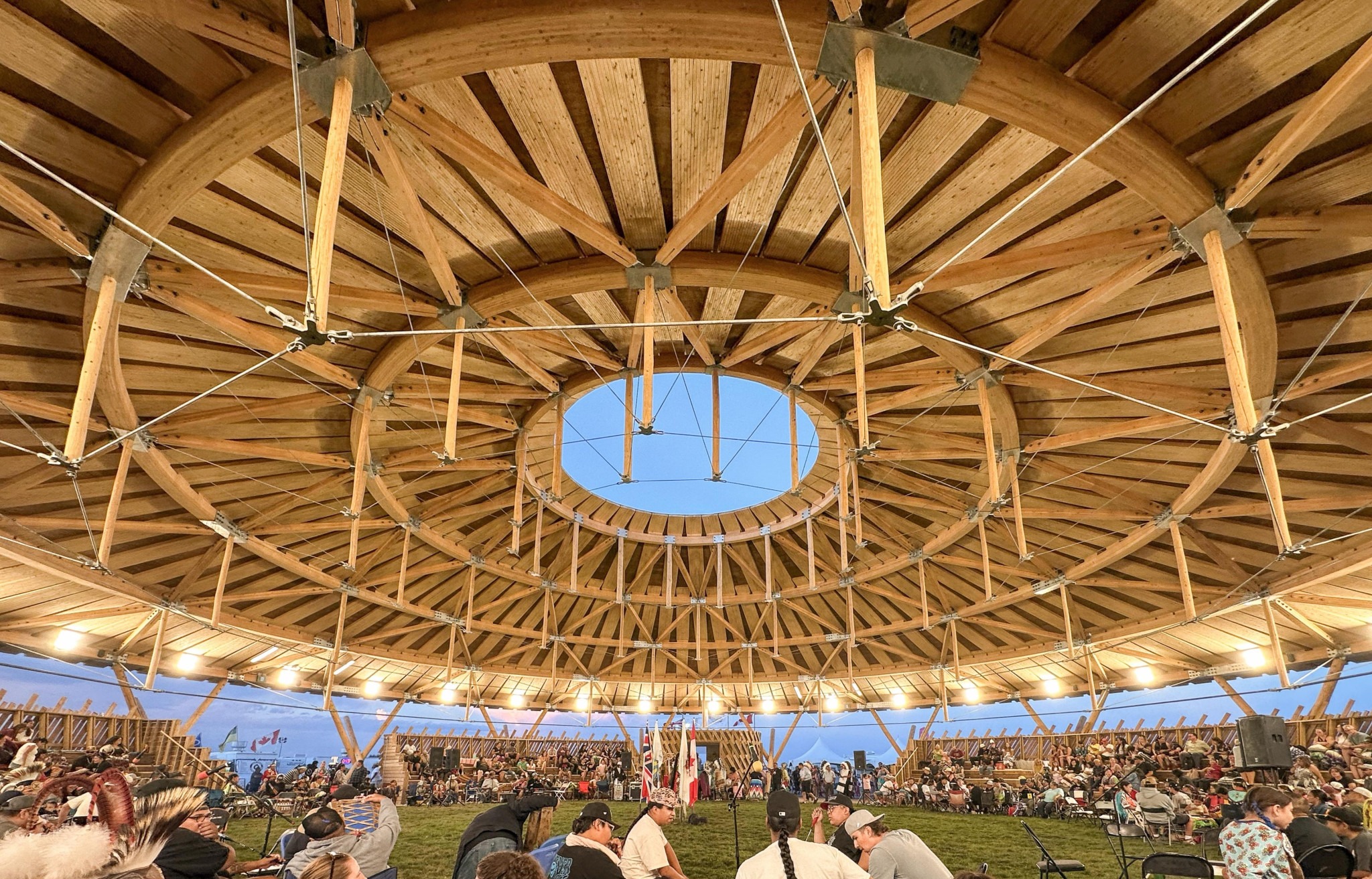 Circular wooden pavilion roof with a skylight, overlooking an event. People gather on grass, creating a communal, warm atmosphere.