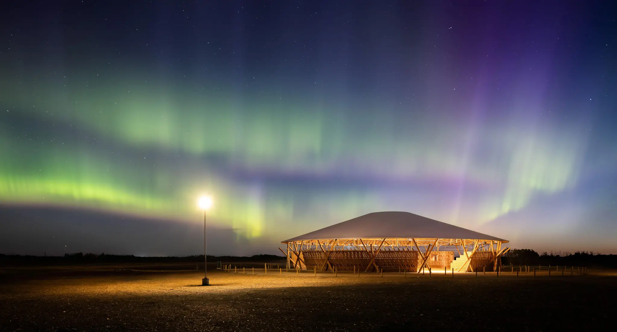 A serene night scene with vibrant green and purple Northern Lights over a wooden pavilion lit by a single streetlamp, set in a vast open field.