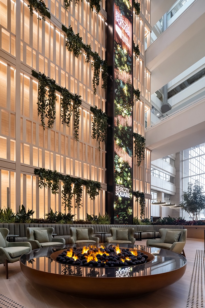 Modern atrium with tall green walls, cascading plants, and warm lighting. A round fire pit is surrounded by plush seating, creating a cozy ambiance.