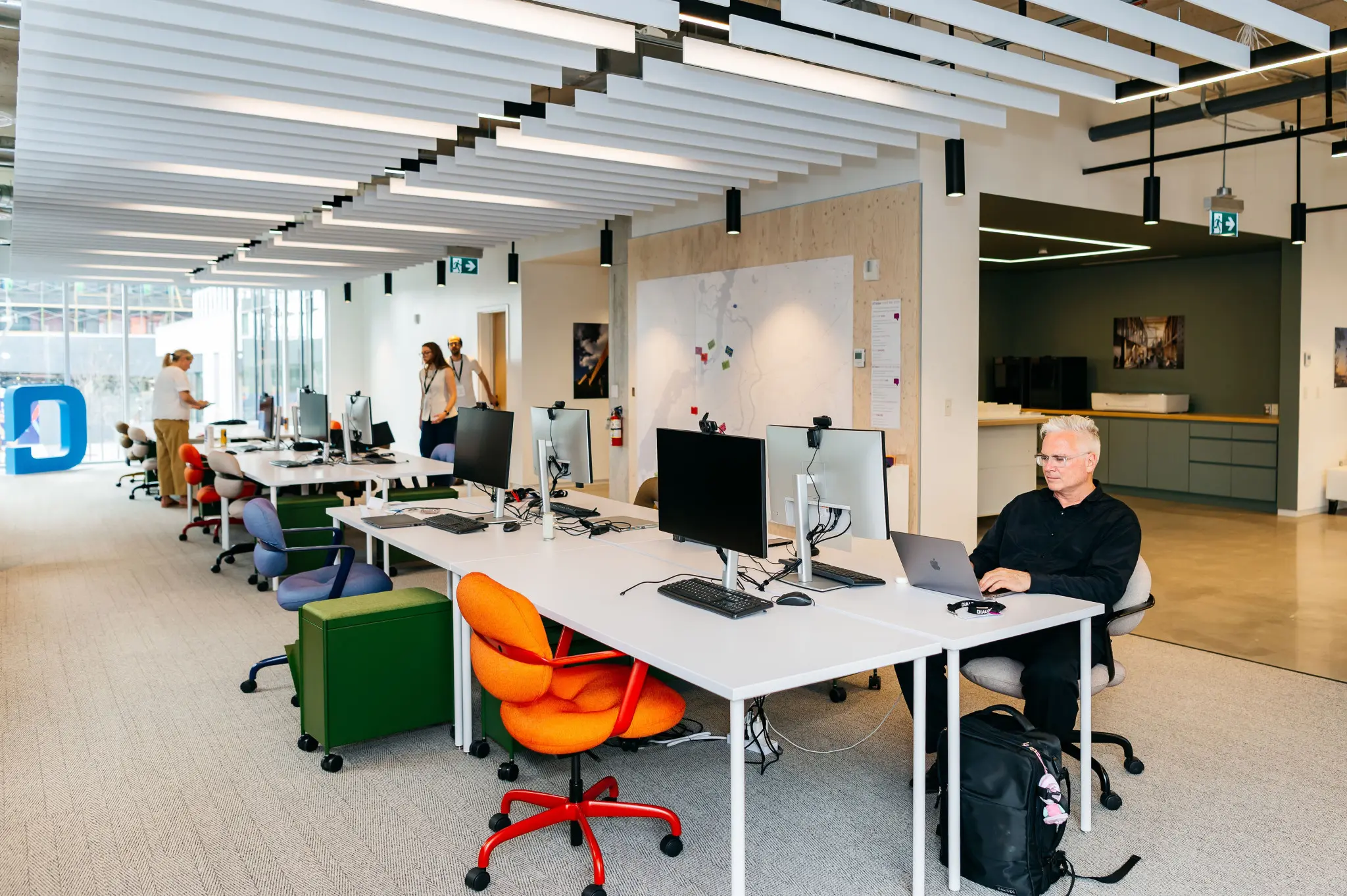 Modern office with open layout and colorful chairs. One person works on a laptop, while others converse. The atmosphere is bright and collaborative.