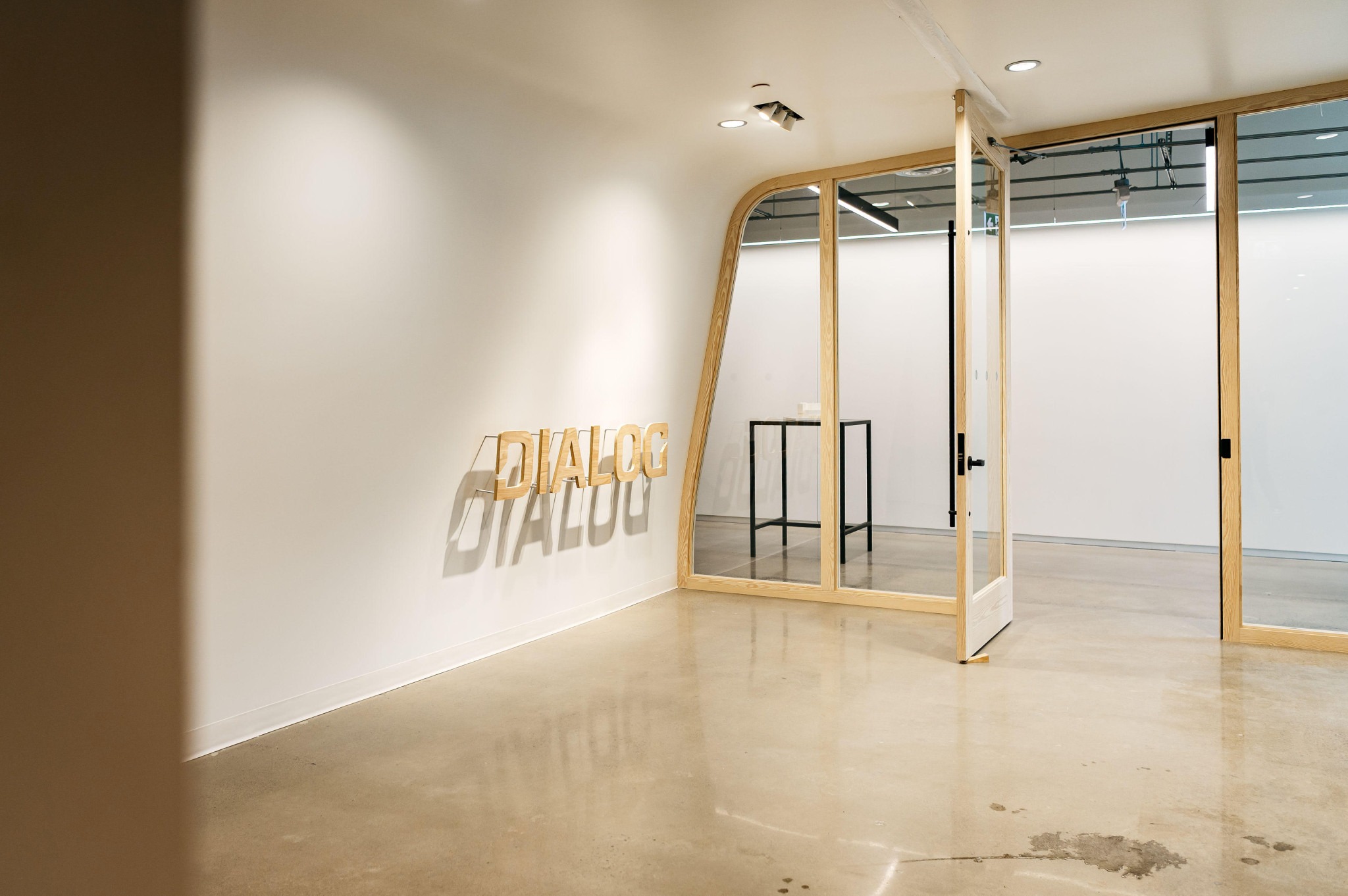 Modern interior with polished concrete floors, a wooden-framed glass door, and the word "DIALOG" casting a shadow on a white wall, creating a minimalist and calm atmosphere.