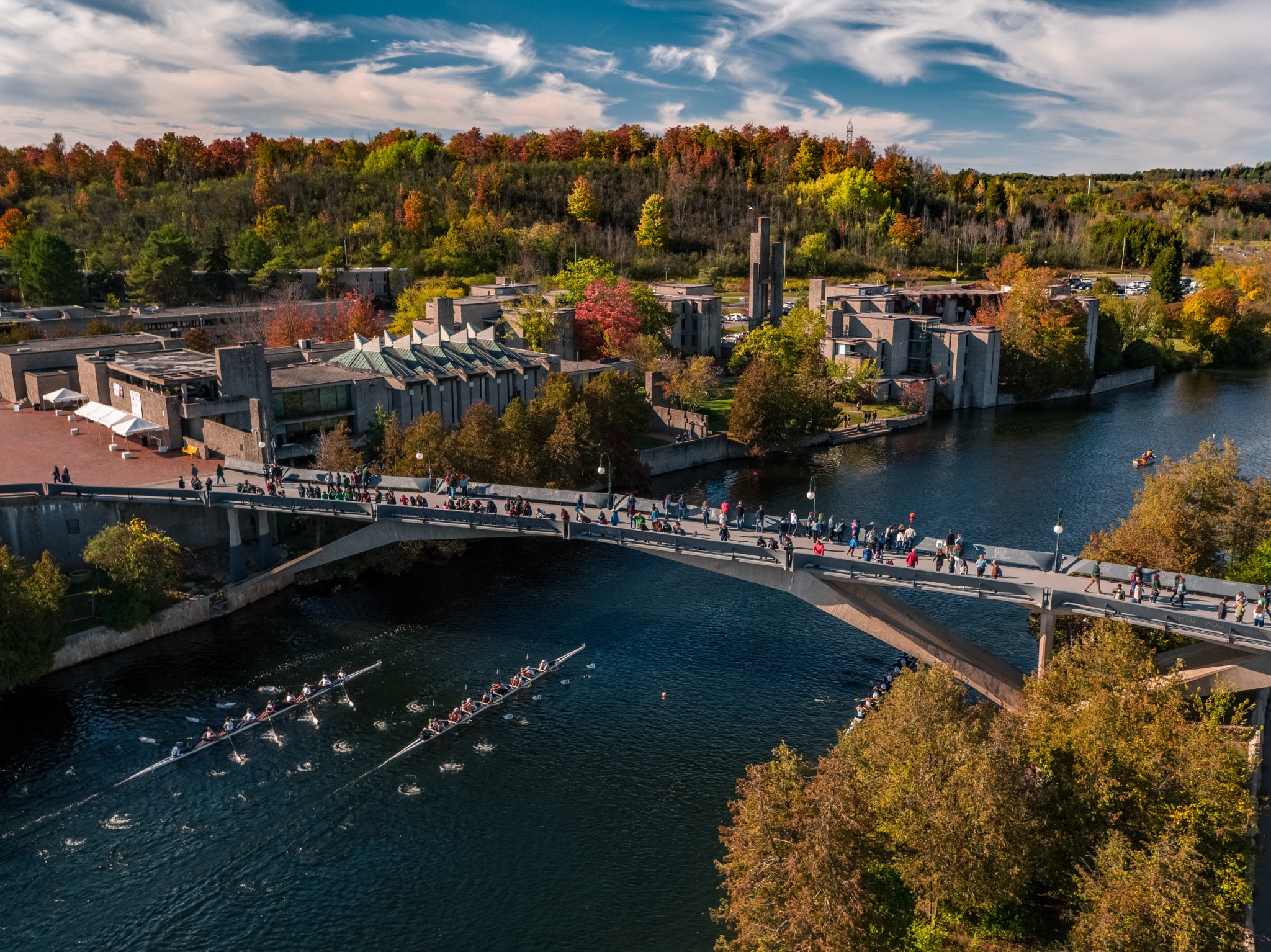 Aerial view of a scenic bridge crowded with people over a tranquil river, with rowers below and surrounded by autumn foliage and rustic buildings.