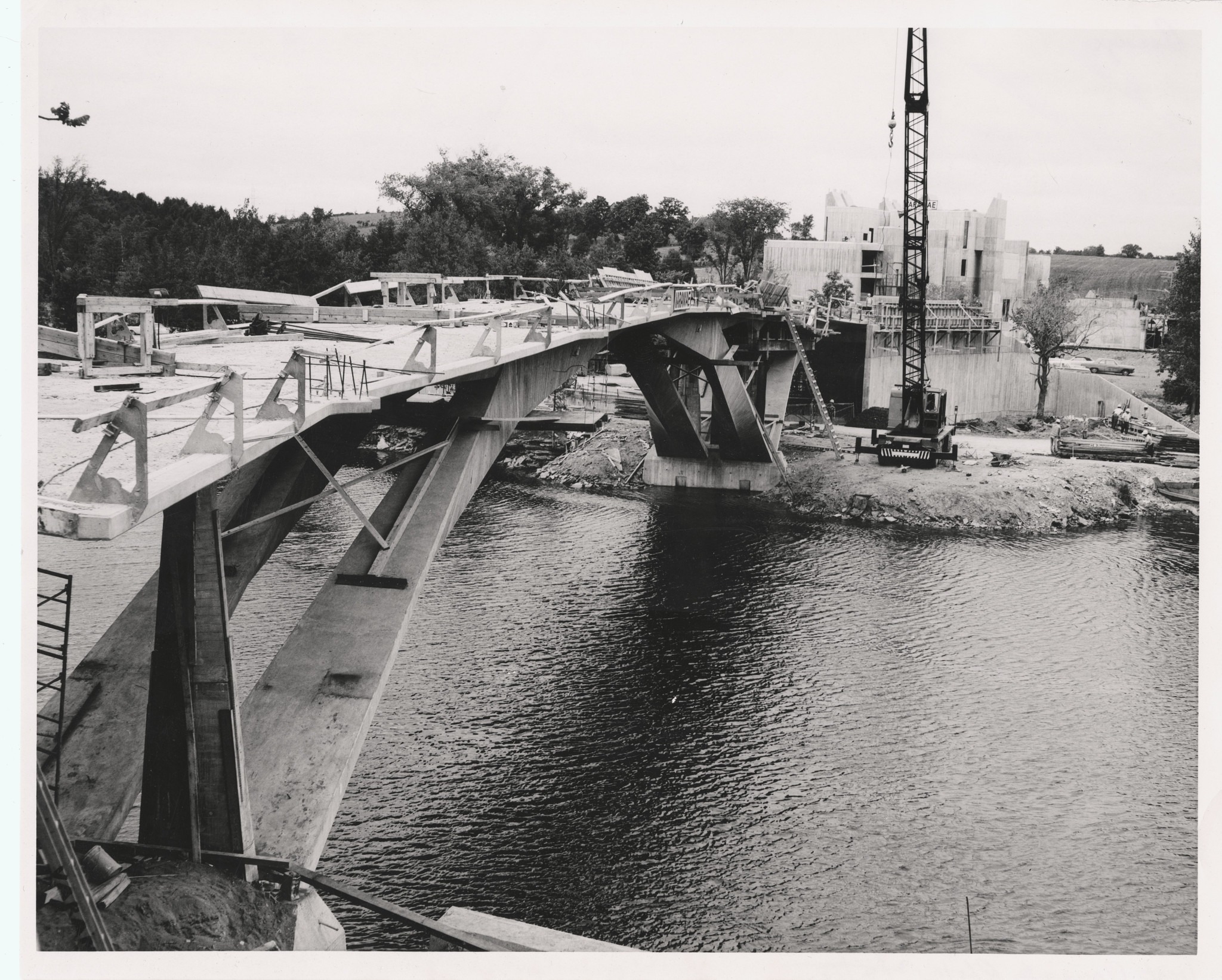 A partially constructed concrete bridge spans a river. A crane and scaffolding are visible amid construction materials. Trees line the riverbank.