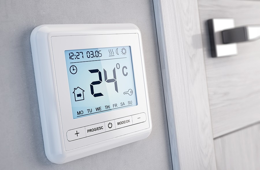 A digital thermostat set to 24°C is mounted on a light gray wall near a wooden door with a silver handle. It displays time, date, and weekday symbols.