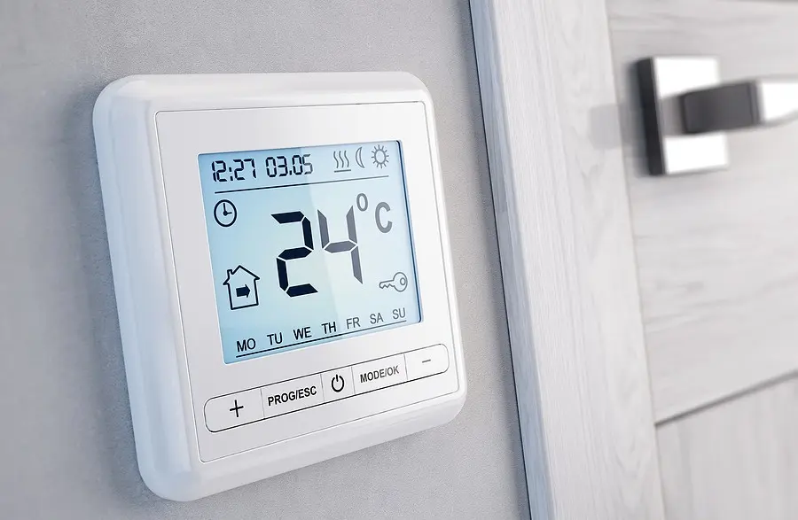 A digital thermostat set to 24°C is mounted on a light gray wall near a wooden door with a silver handle. It displays time, date, and weekday symbols.