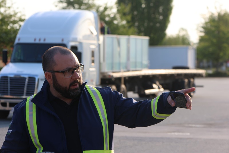 A man in a reflective safety jacket gestures with his hand in a parking lot. A large white truck and flatbed trailer are in the background.