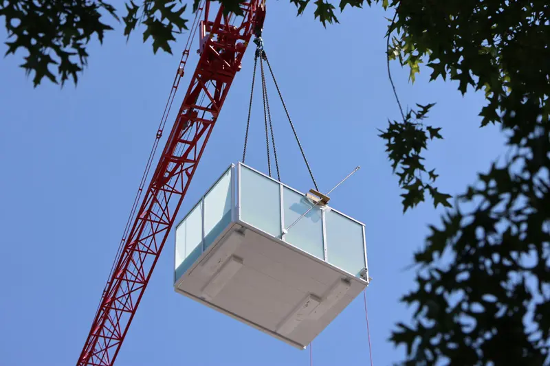 A red crane lifts a glass balcony against a clear blue sky, framed by tree leaves. The scene conveys construction and modern architectural development.