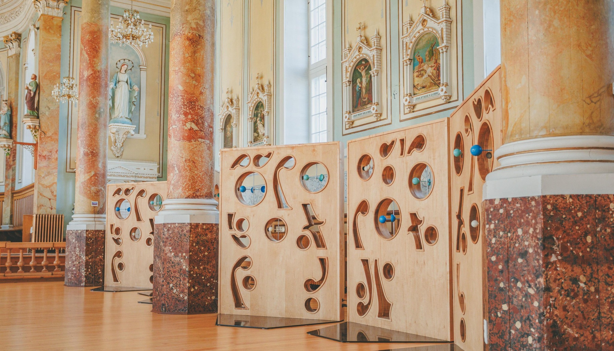 Wooden panels with abstract cutouts and blue accents stand in a grand hall with ornate columns and religious statues, creating an artistic contrast.