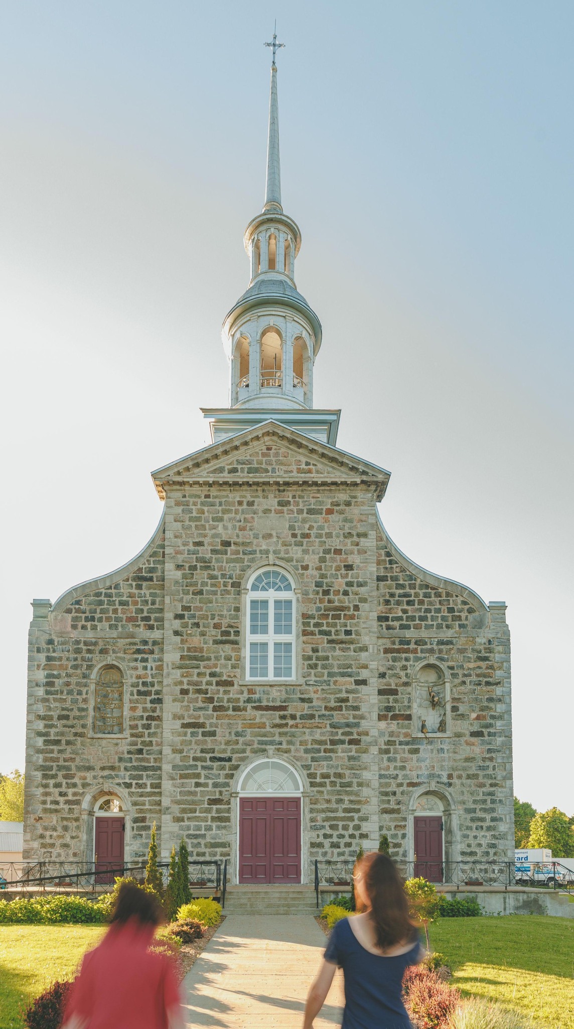 Two blurred figures walk toward a historic stone church with a tall steeple and red doors, set against a clear blue sky, conveying a serene and timeless atmosphere.