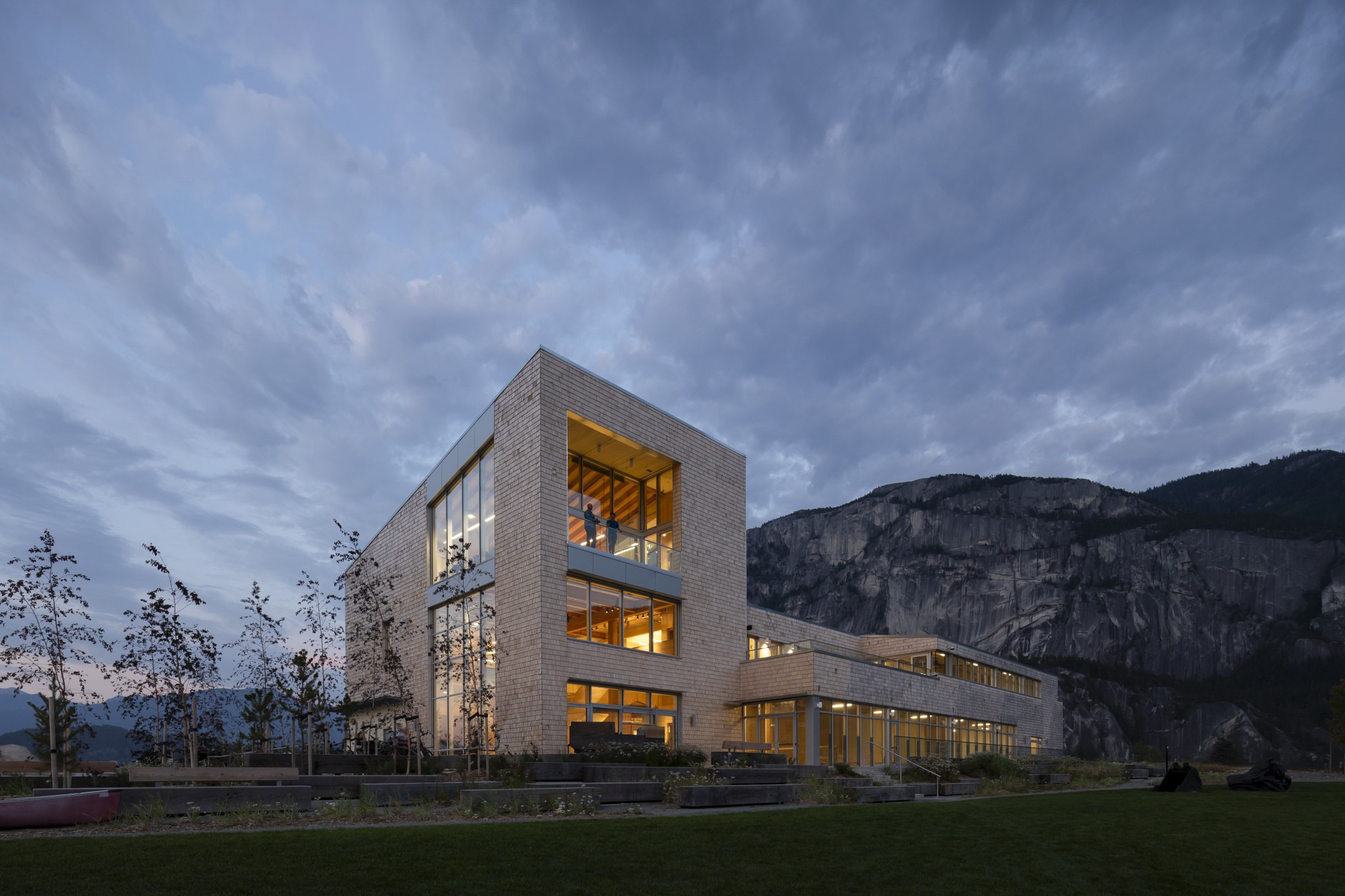A modern building with large glass windows glows warmly at dusk, set against a backdrop of rugged mountains and a cloudy sky.