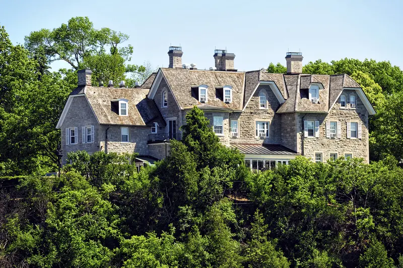 A large, elegant stone mansion with multiple chimneys and windowed dormers is surrounded by lush green trees under a clear blue sky, conveying tranquility.