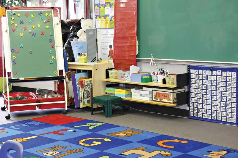 A colorful classroom with a blue carpet featuring letters, a movable easel with magnetic letters, a shelf with educational materials, and a chalkboard on the wall.