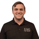 Smiling man in a black polo shirt with "UMI, The Source" embroidered on the chest. White background. Confident and approachable expression.