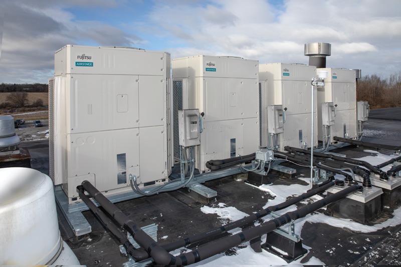 Rooftop shot of four Fujitsu Airstage HVAC units on a flat surface, with connected pipes. Background of cloudy sky and distant trees. Cold, industrial setting.