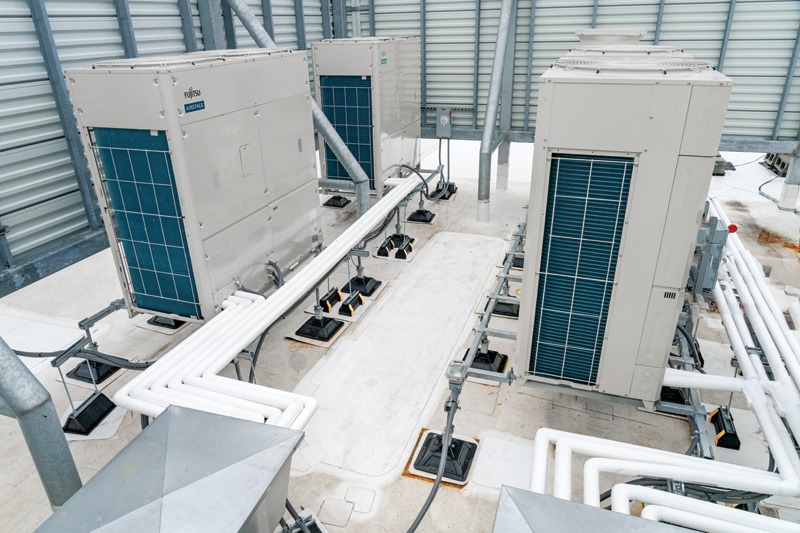 Rooftop view of multiple large air conditioning units with connected white pipes. The setting is industrial, featuring a clean, organized layout.