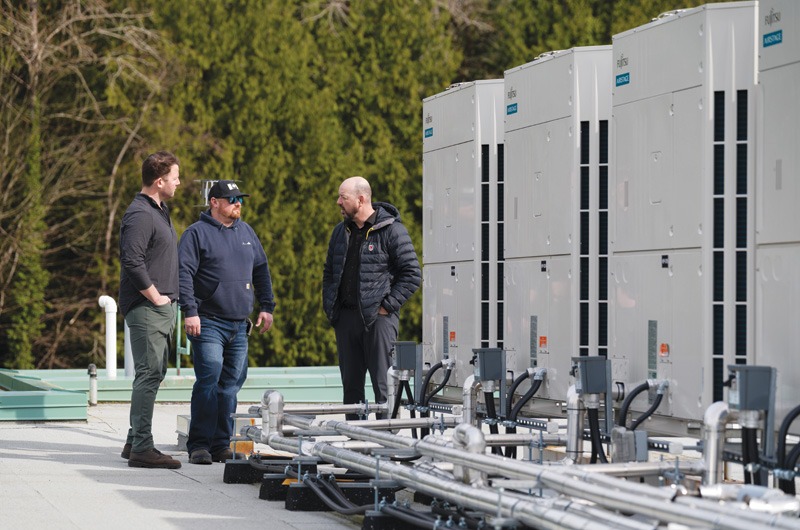 Three men stand on a rooftop beside large HVAC units, discussing. They wear casual clothing and jackets. Trees are visible in the background.