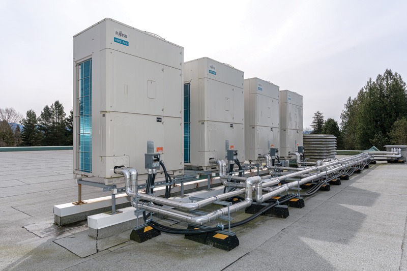 Rooftop HVAC units with connected pipes on a flat roof. Overcast sky, trees in the background; industrial and organized setting.