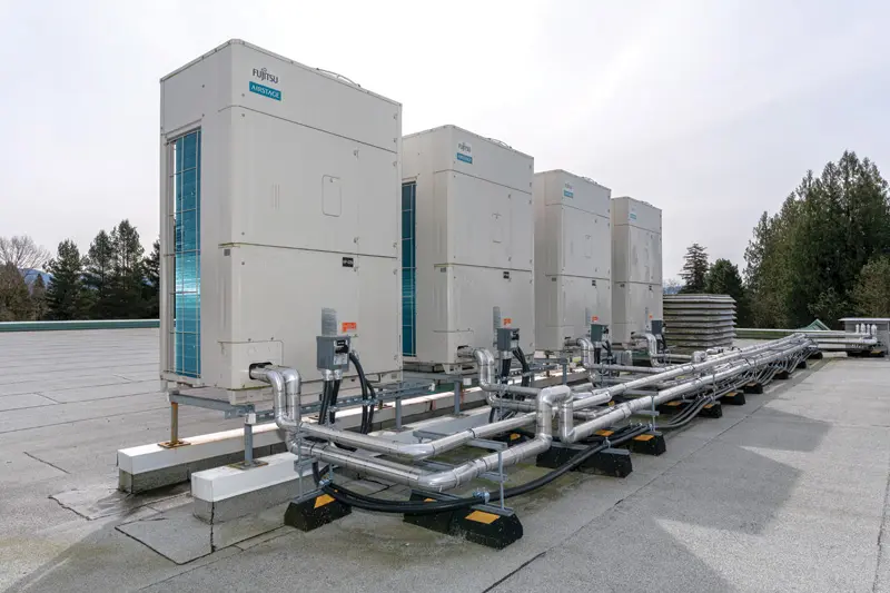 Rooftop HVAC units with connected pipes on a flat roof. Overcast sky, trees in the background; industrial and organized setting.