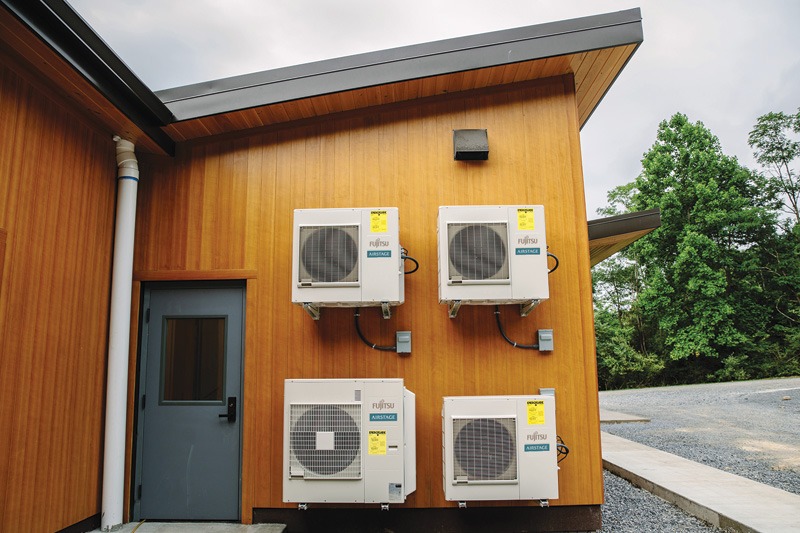 Wooden building exterior with four mounted air conditioning units on the wall near a gray door. Lush green trees in the background under a cloudy sky.