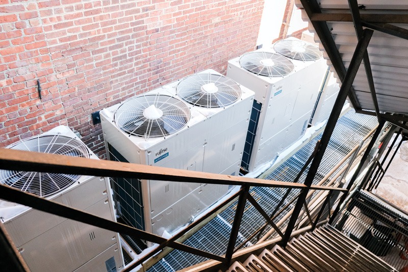 Four large industrial air conditioning units lined up against a brick wall, viewed from above a metal staircase, creating an orderly, functional scene.