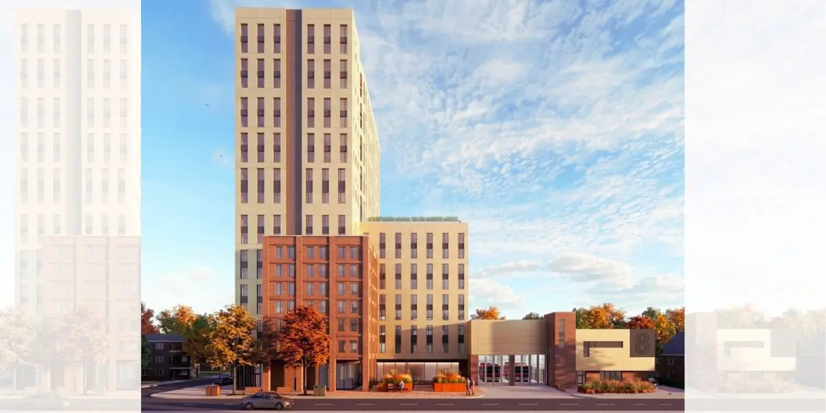 A modern multi-story building with a light beige tall section and a red brick base, surrounded by autumn-colored trees under a blue sky.