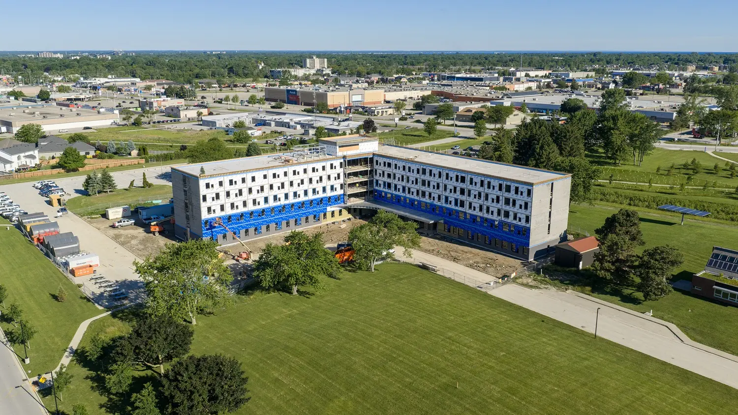 Aerial view of a large, U-shaped building under construction in a suburban area. Surrounded by greenery and parking lots, it conveys a sense of growth.